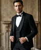 Man wearing a black tuxedo with a bow tie against a stone wall background
