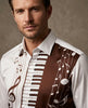 A close-up of a man wearing a white button-up shirt with a brown and white musical-themed print featuring piano keys, musical notes, and a treble clef.