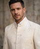 A man wearing a cream-colored embroidered Indian sherwani jacket over a white collared shirt, set against a blurred stone wall background.