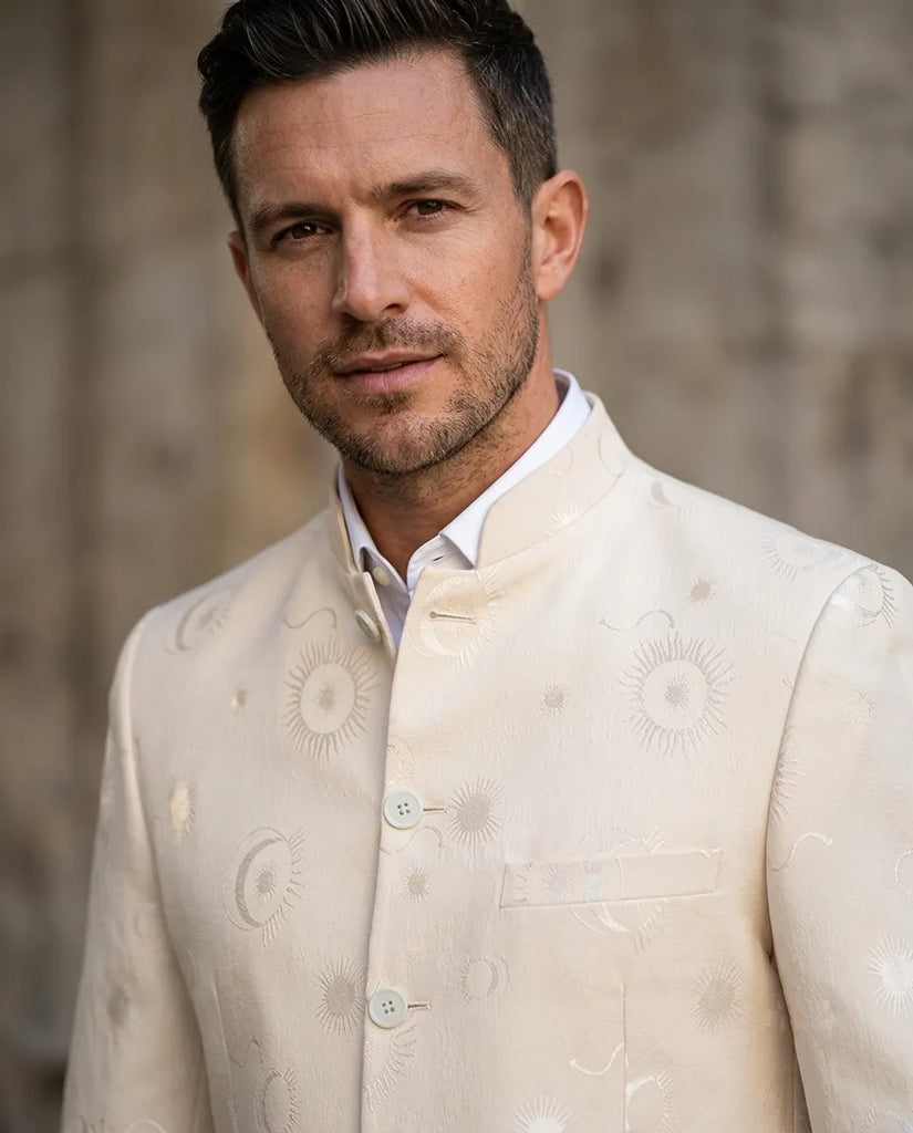 A man wearing a cream-colored embroidered Indian sherwani jacket over a white collared shirt, set against a blurred stone wall background.