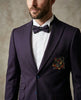 A man wearing a dark purple suit jacket with a white dress shirt, a matching dark purple bow tie, and a multicolor embroidered pocket square on the breast pocket, set against a textured neutral background.