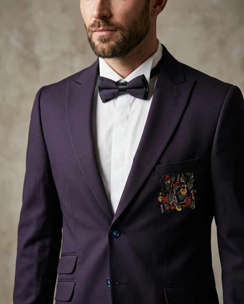 A man wearing a dark purple suit jacket with a white dress shirt, a matching dark purple bow tie, and a multicolor embroidered pocket square on the breast pocket, set against a textured neutral background.