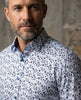 Man wearing a white shirt with a subtle blue floral pattern against a textured wall.