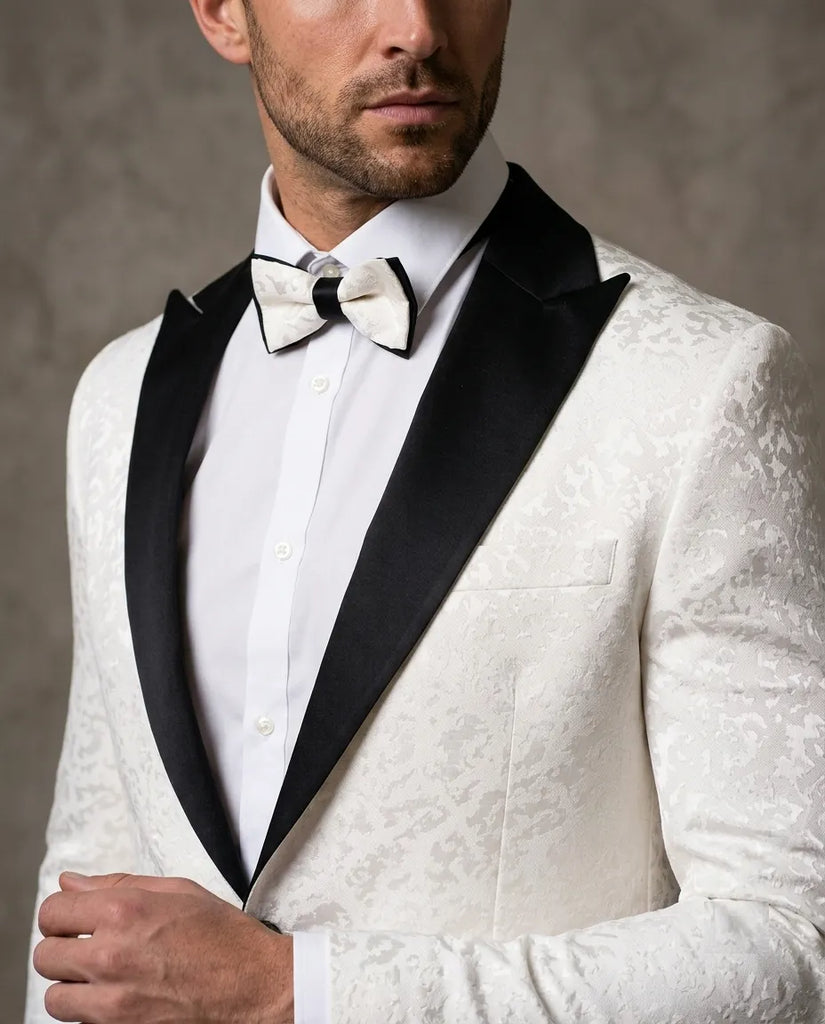 A man wearing a white patterned tuxedo jacket with black satin lapels, a white dress shirt, and a white bow tie with black accents, shown in a close-up studio portrait.