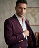 A man wearing a deep burgundy double-breasted blazer with gold buttons over a white dress shirt, posed in a warm, softly lit indoor setting.