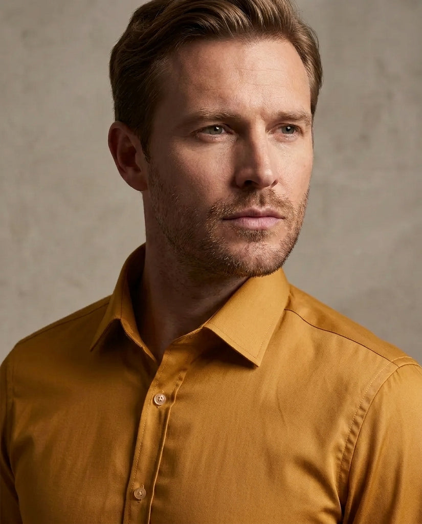 A close-up portrait of a man wearing a mustard yellow button-down shirt against a textured gray background.