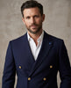 A man wearing a navy blue double-breasted suit jacket with gold buttons, styled over a white dress shirt, photographed against a textured neutral background.